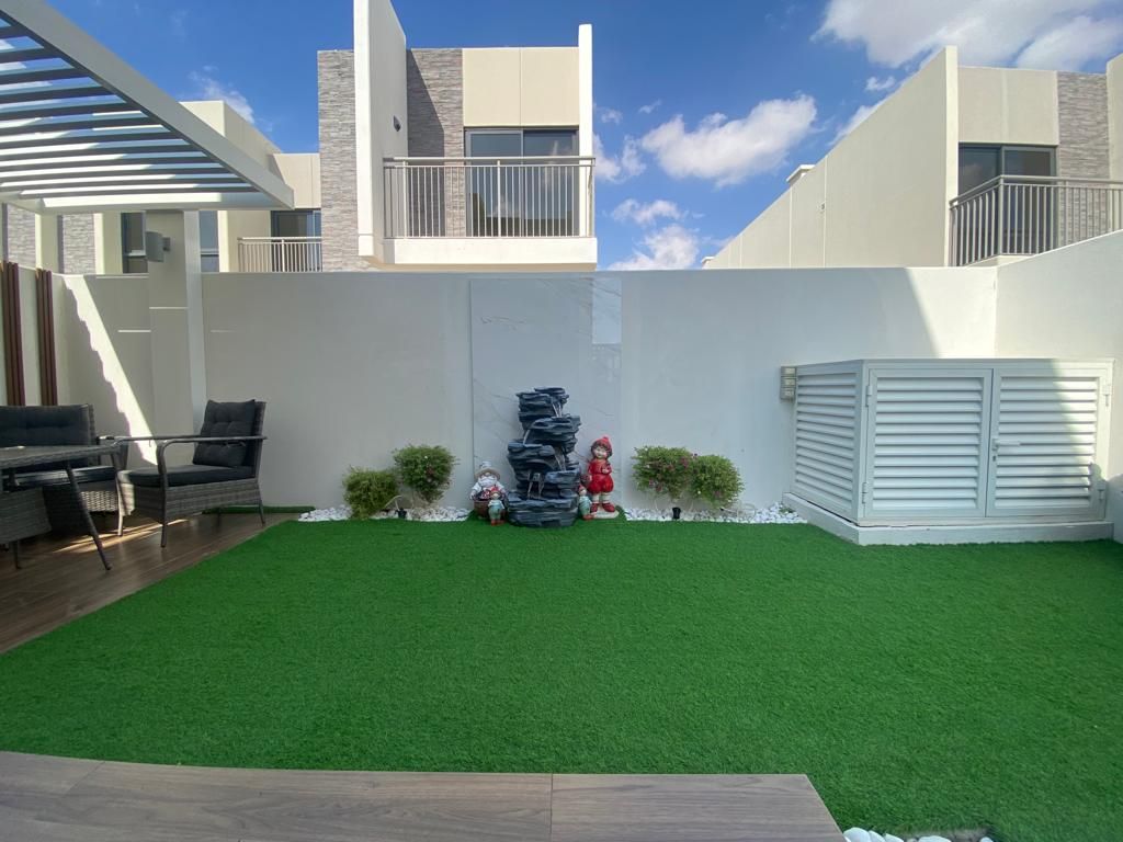 Lawn of artificial grass beside a wood-look deck with wicker-look outdoor seating, facing a rock-style water feature, small flowering shrubs and white pebbles along the white boundary wall, Project Alpha, Damac Hills 2