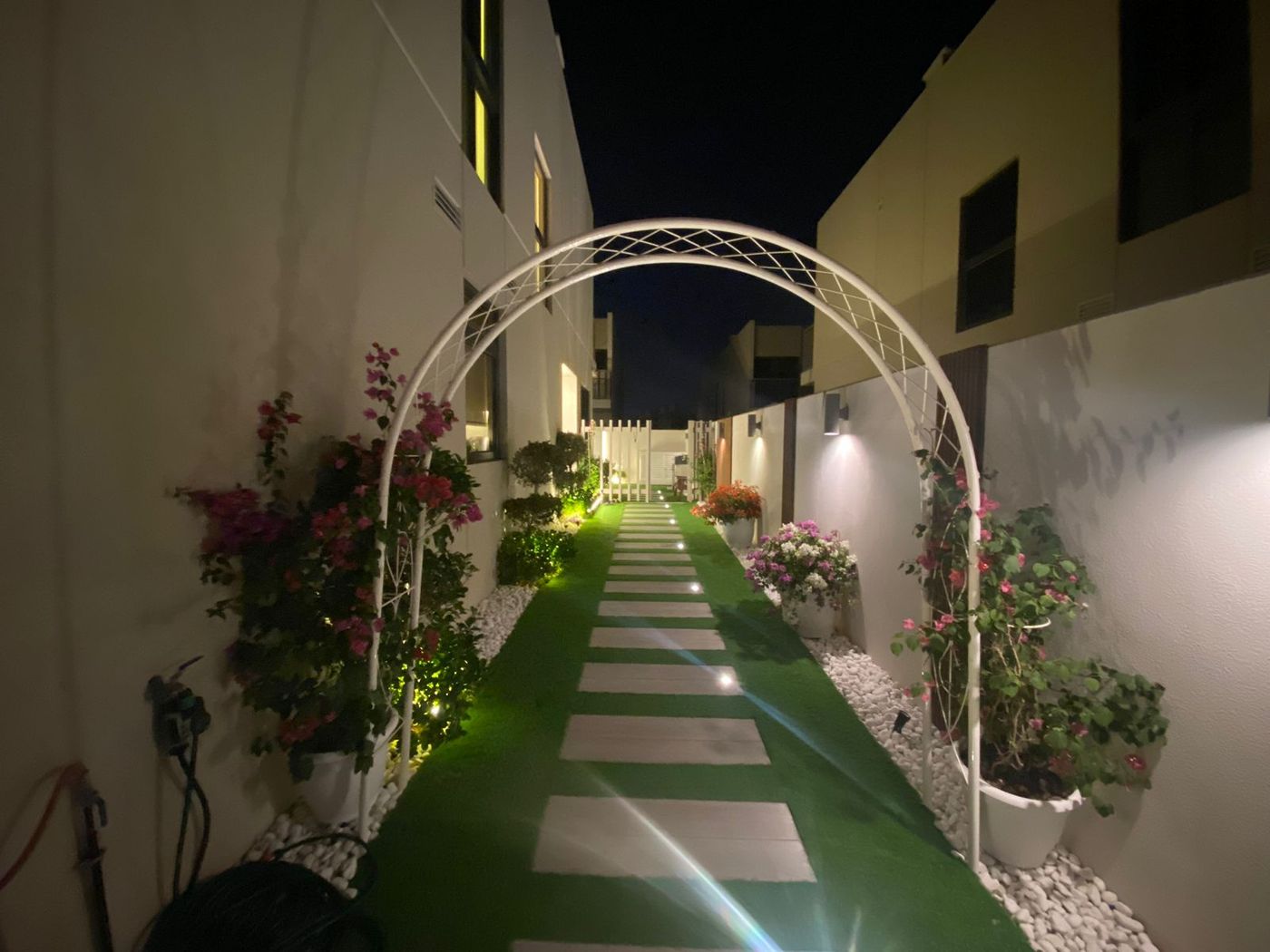 Side passage at night with a white garden arch over stepping stones set in artificial grass, potted flowering plants on both sides, in-ground lights and wall lights