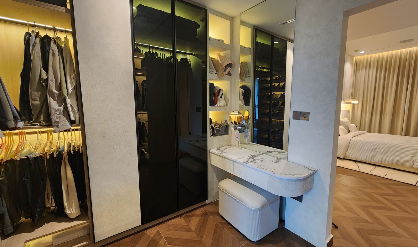 Dressing area opening onto a bedroom: open hanging rails, smoked-glass wardrobe doors and a dressing table with a marble-look top and a stool, on a chevron-pattern floor