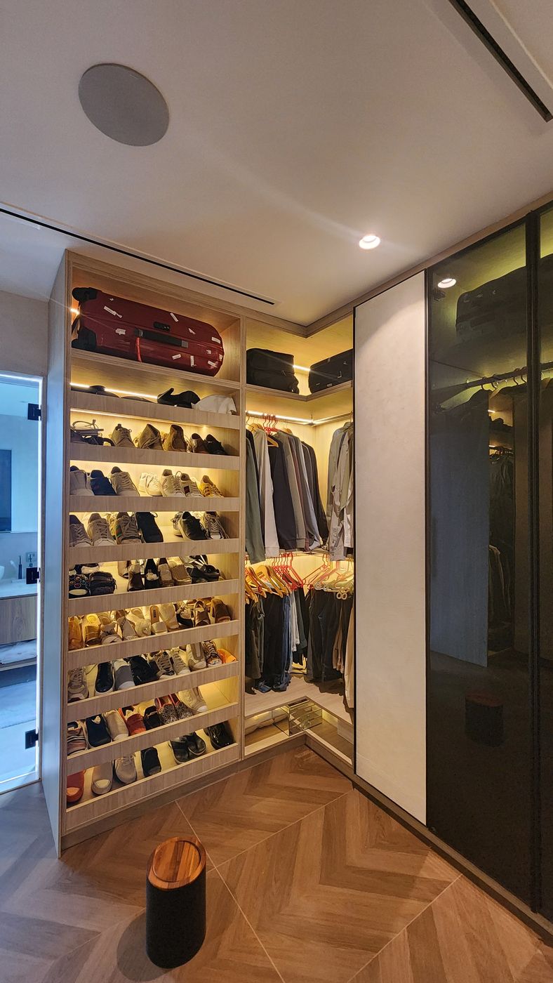 Corner of a dressing area with lit, angled shoe shelves, corner hanging rails, suitcases on the top shelves and smoked-glass wardrobe doors