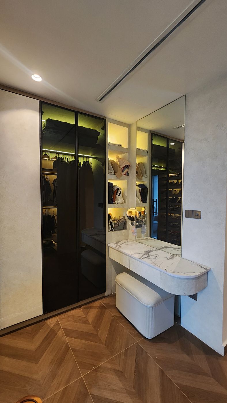Tall smoked-glass wardrobe doors next to a dressing table with a marble-look top, an upholstered stool, lit open shelves and a tall wall mirror