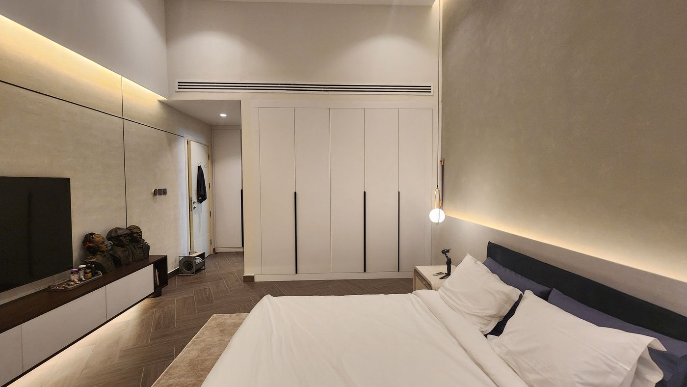 Wide view of a bedroom with a run of white fitted wardrobes with black bar handles, a wall-mounted TV above a low unit lit from underneath, and a made bed