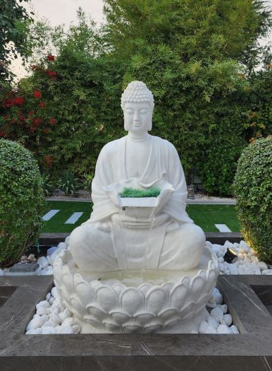 White seated Buddha statue pouring water from a bowl into a lotus-shaped basin set in white pebbles, framed by round hedges