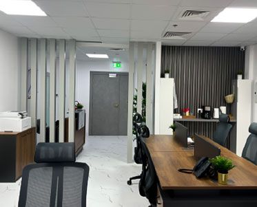 Open-plan office with wood-tone desks and black mesh chairs behind light grey vertical slat partitions, a grey door with an exit sign, marble-look floor tiles and a dark slatted coffee corner at the right, Project Door Step, Silicon Oasis