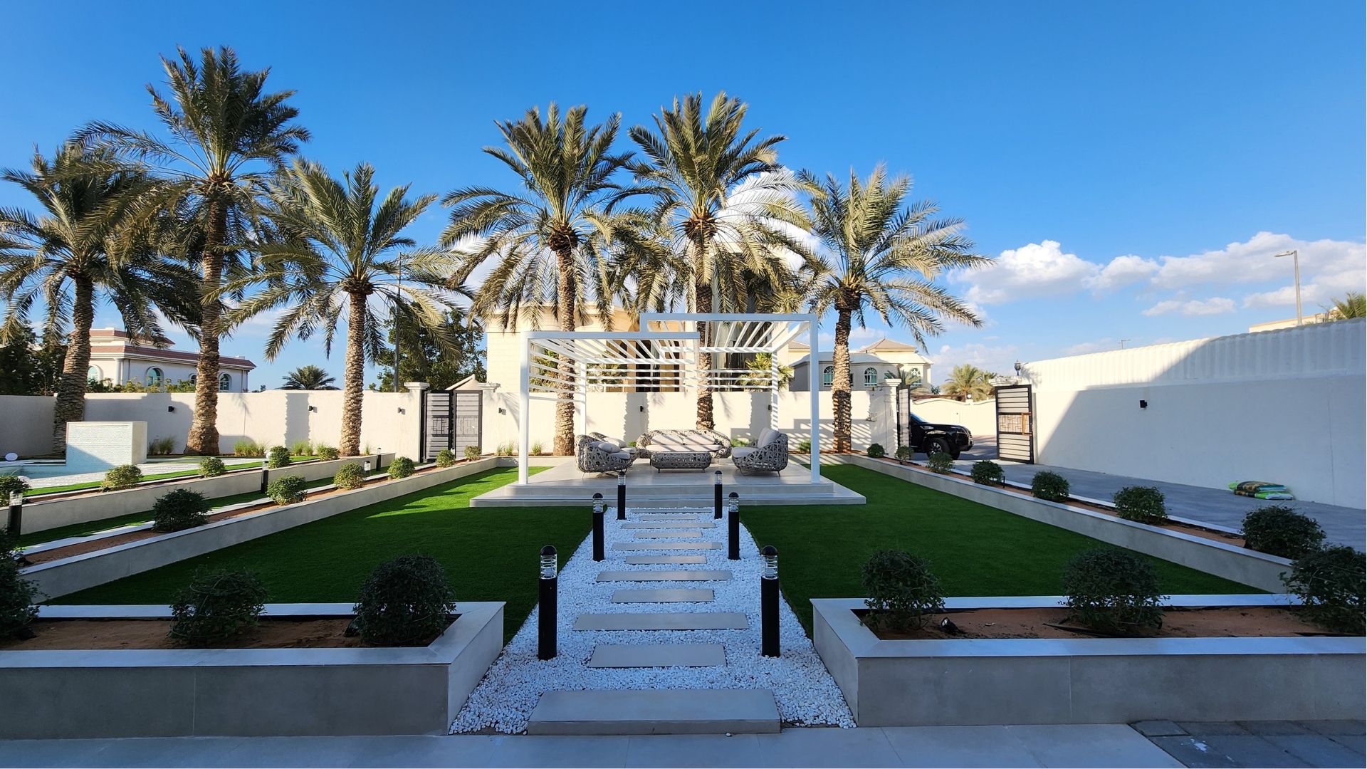 Wide view of a finished villa garden with raised planters, a white stone path, gazebo and date palms, Project Majestic, Al Twar