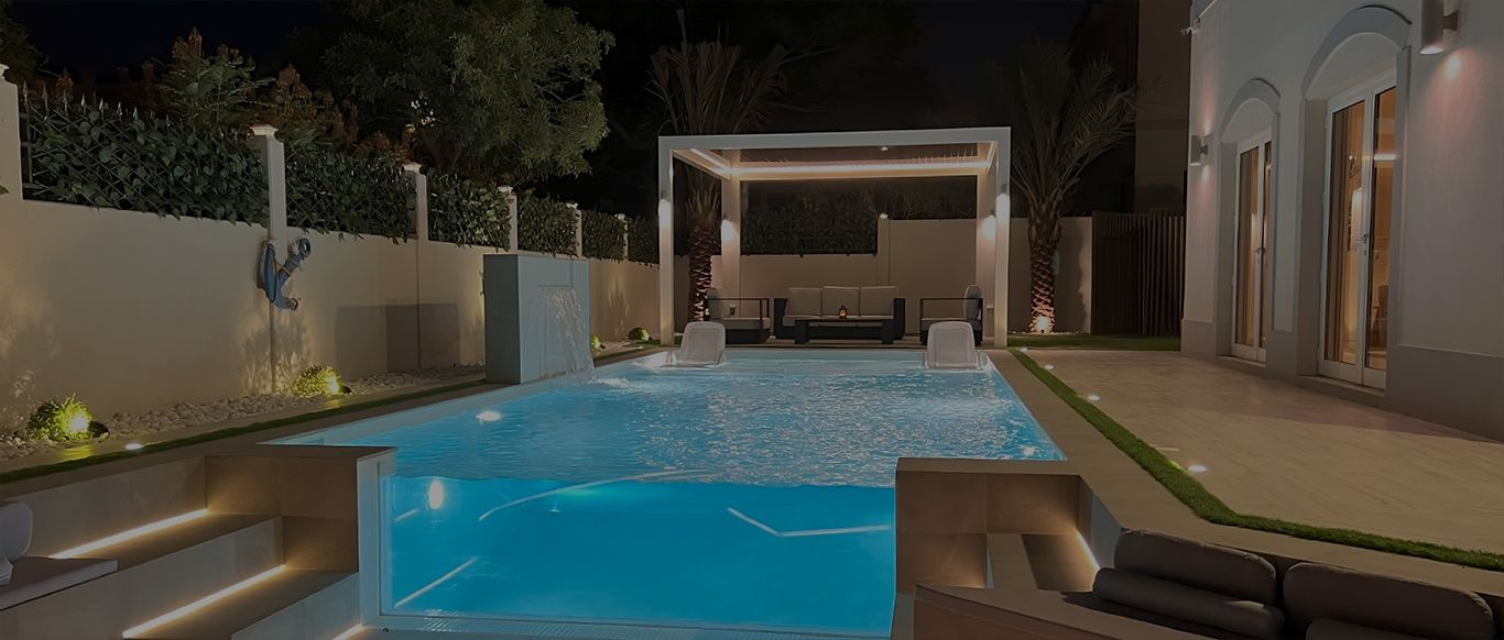 Lit pool at night with a glass corner panel, a sheet waterfall falling from a raised block and two loungers in the water, with an outdoor sofa set under a white louvred roof beyond, Project Miracle, The Villa