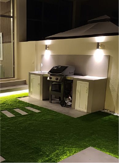 Built-in BBQ counter with cabinets, a backsplash and a BBQ station, lit by wall lights, beside artificial grass at night, Project Murano, Al Furjan