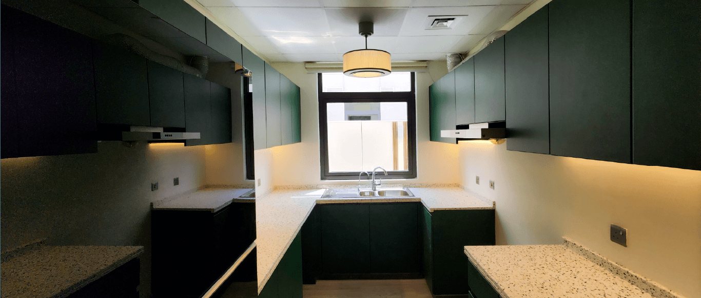 U-shaped kitchen with dark green flat-front cabinets, light speckled worktops, warm lighting under the wall units and a drum pendant in front of the window over a double sink, Project Solace, DAMAC Hills