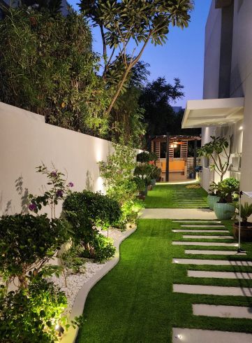 Narrow side garden at dusk with curved planting beds edged in white pebbles, wall uplights and stepping stones in grass