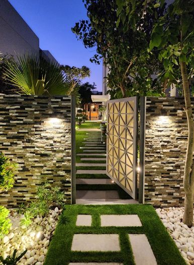 Open garden gate with a geometric pattern between two stone-look clad walls with wall lights, stepping stones in grass in front